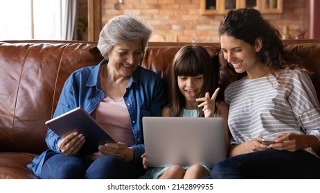 Happy Kid Girl, Young Mother, Older Grandmother Using Digital Devices, Laptop, Tablet, Making Video Call, Watching Movie Online, Reading Book. Three Family Female Generations Using Electronic Gadgets