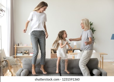 Happy kid granddaughter, mother and grandmother having fun jumping on sofa, cheerful three generations women family dancing in living room, grandma, mom and child laughing spending time together