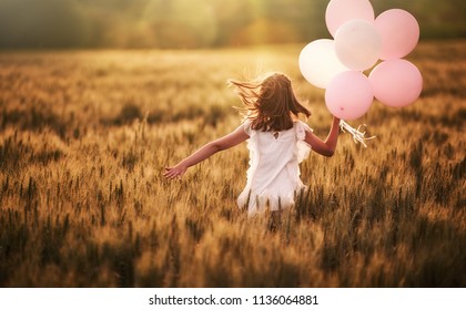 Happy kid is having fun on nature in the summer. Child is playing on meadow at sunset background. Girl with air balloons is running on cereal field.