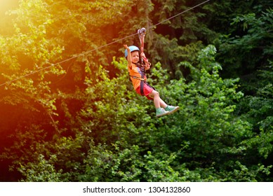 Happy Kid With Helmet And Harness On Zip Line Between Trees