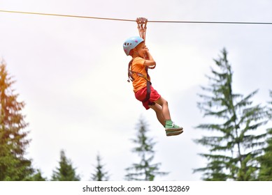 Happy Kid With Helmet And Harness On Zip Line Between Trees