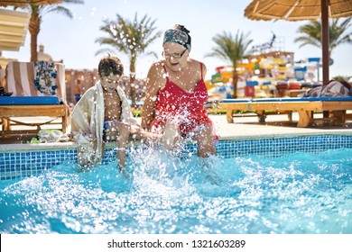 Happy kid with his grandma are shaking their feet in the water while sitting on the edge of the pool on the sunny background of the hotel. Lady wears a red swimsuit with headband and glasses.