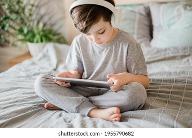 Happy Kid Lying On The Bed At Home And Playing With Pc Tablet Or Reading Online And Listening To Music With Wireless Headphones