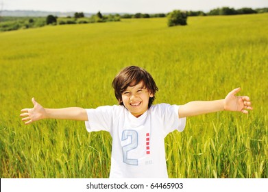 Happy Kid In Nature, Positive Smiling Child On Green Beautiful Meadow With Open Arms Looking In Camera