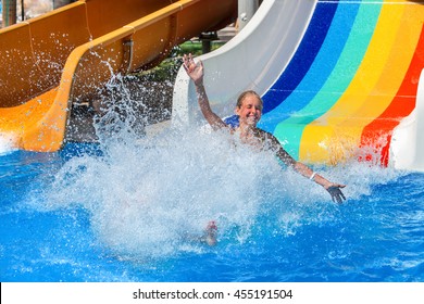 Happy Kid On Water Slide At Water Park Hands Up. There Are Some Water Slides With Flowing Splash Water In Aqua Park. Summer Water Park Holiday. Outdoor.