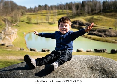 Happy kid with open arms pretending to be a plane pilot sitting on boulder by a lake is engaging in a funny outdoor activity. Smiling child sitting like an airplane enjoying close contact with nature - Shutterstock ID 2227879715