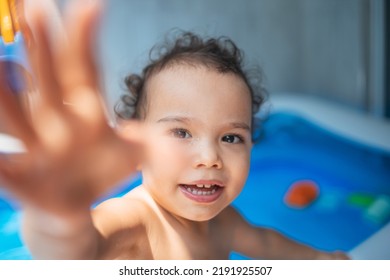 Happy Kid Playing In An Inflatable Pool In Backyard. Curly Toddler Smiling And Playing With Water