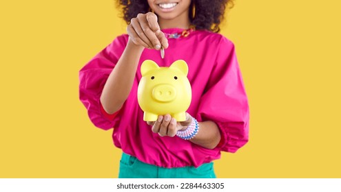 Niño feliz ahorrando algo de dinero. Niña pequeña sosteniendo un banco de cerditos amarillos. Una alegre niña afroamericana pone una moneda dentro de su pequeño lechón. Recortada. Fondo del cartel. Concepto de finanzas