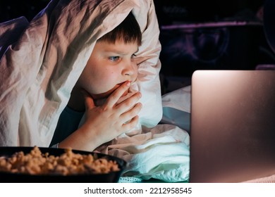 Happy Kid Son Laughing Eat Popcorn Remote Control Watching Funny Comedy Tv Show Sitting On Sofa Having Fun Viewing Video On Laptop In Evening At Home. Teenager Gen Z Boy Under Blanket At Movie Night