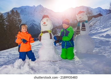 Happy Kids Build, Dress Snowman With Scarf And Hat Outside Over Mountain Peaks On Background