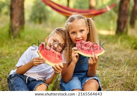 Happy Kids eating watermelon in the park. Kids eat fruit outdoors. Healthy snack for children. Little girl and boy playing in the forest biting a slice of water melon