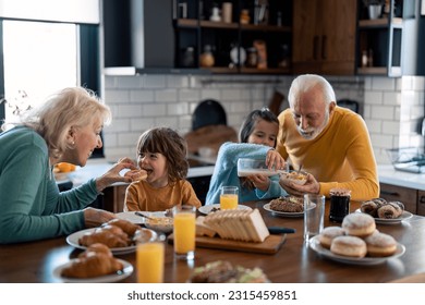 Happy kids enjoying delicious breakfast with caring proud grandparents. Little girl pouring milk into cornflakes cereals with grandpas help while grandma is feeding her little brother with donut. - Powered by Shutterstock