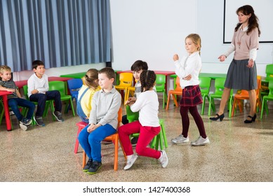 Happy Kids And Female Teacher Playing Musical Chairs Together During Break In Classroom At Elementary School