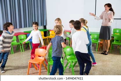 Happy Kids And Female Teacher Playing Musical Chairs Together During Break In Classroom At Elementary School