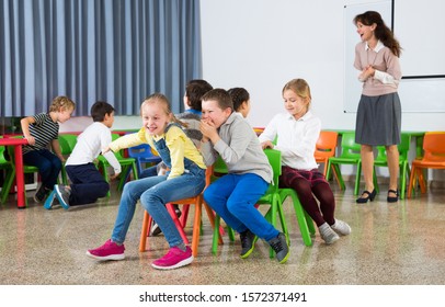 Happy Kids And Female Teacher Playing Musical Chairs Together During Break In Classroom At Elementary School