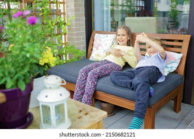 Happy Kids Have Fun And Rest On Cozy Balcony At Home. Wooden Table, Many Green Plants, Soft Sofa, Candles. Lovely Terrace Life On Vacation. Green Place In City Apartment