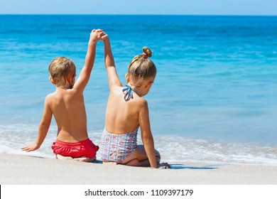 Happy kids have fun in sea surf on white sand beach. Couple of children sit in water pool with hands up. Travel lifestyle, swimming activities in family summer camp. Vacations on tropical island.