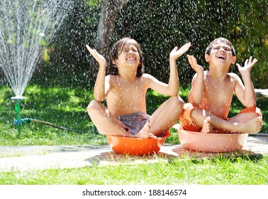 Happy Kids Playing And Splashing With Water Sprinkler On Summer Grass Yard