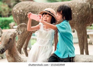 Happy Kids Playing And Take Photo By Smartphone, Family Children Holding Mobilephone And Smiling, Asain Child Sitting On Deer Statue In Zoo And Learning Outdoors On Summer Holiday 