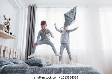 Happy Kids Playing In White Bedroom. Little Boy And Girl, Brother And Sister Play On The Bed Wearing Pajamas. Family At Home