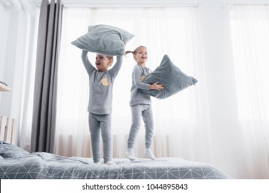 Happy Kids Playing In White Bedroom. Little Boy And Girl, Brother And Sister Play On The Bed Wearing Pajamas. Family At Home