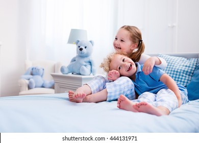 Happy Kids Playing In White Bedroom. Little Boy And Girl, Brother And Sister Play On The Bed Wearing Pajamas. Nursery Interior For Children. Nightwear And Bedding For Baby And Toddler. Family At Home.