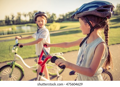 Happy Kids Are Riding Bikes Outdoors, Looking At Each Other And Smiling