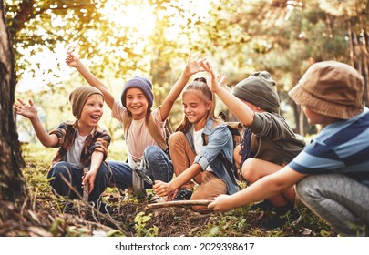Happy kids scouts learning how to create fire with magnifying glass and sun in forest during school camping trip, screaming in excitement while squatting together in nature near pile of dry branches