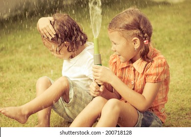 Happy Kids Sitting On The Grass And Pouring Water From A Hose. Concept Brother And Sister Together Forever