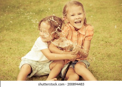 Happy Kids Sitting On The Grass And Pouring Water From A Hose. Concept Brother And Sister Together Forever