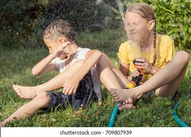 Happy Kids Sitting On The Grass And Pouring Water From A Hose. Concept Brother And Sister Together Forever.