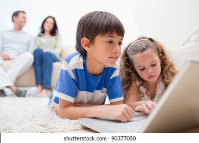 Happy Kids Using Laptop On The Carpet With Parents Behind Them