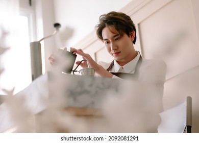 Happy Korean Barista Man Wearing Brown Apron Making Hand-drip Coffee, Pours Hot Water From Black Teapot Into A Paper Filter. Alternative Ways Of Brewing Coffee. Man Barista Cafe Owner Concept