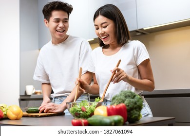 Happy Korean Couple Cooking Together And Laughing Preparing Dinner Standing In Kitchen At Home. Asian Family Making Veggie Salad Indoors. Healthy Nutrition And Recipes Concept