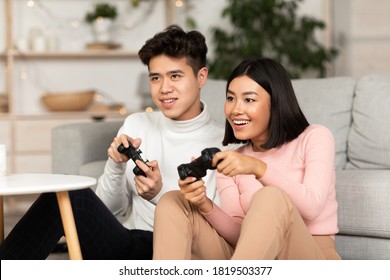 Happy Korean Couple Playing Computer Game Holding Joysticks Sitting On Couch At Home On Weekend. Chinese Boyfriend And Girlfriend Play Video Games. Family Weekend Fun, Computer Gaming Concept