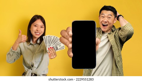 Happy korean couple recommending nice gambling mobile app, showing cell phone with black blank screen and cash dollars on yellow studio background, woman gesturing thumb up, mockup, collage, panorama