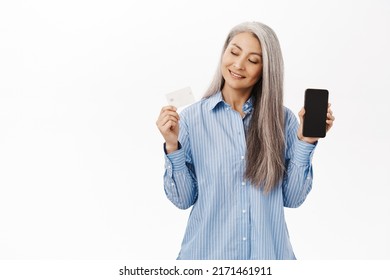 Happy Korean Senior Woman, Grandmother Showing Mobile Phone Screen And Credit Card, Recommending Bank, Online Shopping App, Standing Over White Background