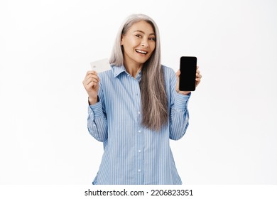 Happy Korean Senior Woman, Grandmother Showing Mobile Phone Screen And Credit Card, Recommending Bank, Online Shopping App, Standing Over White Background.