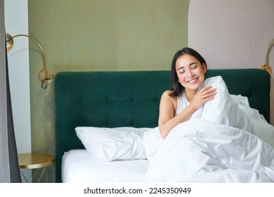 Happy korean woman wakes up in her hotel room, lying in bed under cozy warm blankets, white sheets, enjoying weekend morning. - Powered by Shutterstock