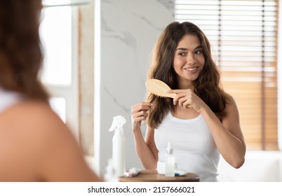 Happy Lady Brushing And Detangling Hair Using Wooden Brush Standing Near Mirror In Modern Bathroom At Home. Hair Beauty And Cosmetics, Treatment And Care. Selective Focus - Powered by Shutterstock