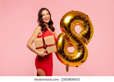 Happy lady celebrating International Women's Day, holding number 8 balloon and wrapped gift box, posing over pink studio background