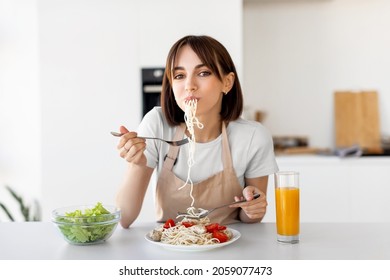 Happy Lady Eating Spaghetti And Vegetable Salad, Enjoying Tasty Lunch, Sitting At Table And Looking At Camera In Modern Kitchen Interior, Copy Space