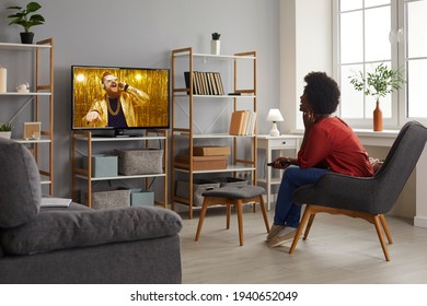 Happy lady enjoying funny TV show sitting in comfy armchair at home. Young woman relaxing in comfortable chair in living-room and watching music video on television