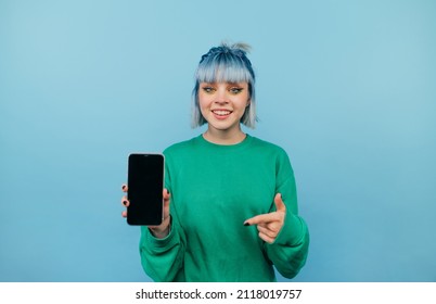 Happy lady in green sweatshirt and colored hair stands on a blue background with a smartphone in his hands and points his finger at a blank black screen.