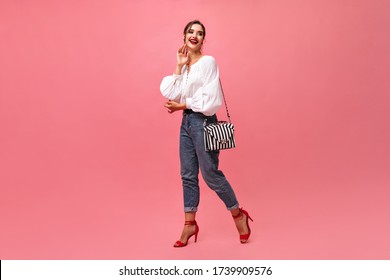 Happy lady in jeans and white blouse posing on pink background. Smiling brunette with red lipstick and in modern outfit moves..