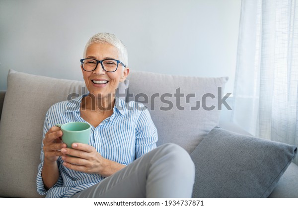 Happy lady relaxing at\
home with hot drink. Shot of a happy mature woman relaxing on the\
sofa with a warm beverage at home. Senior woman drinking hot drink\
sitting on sofa
