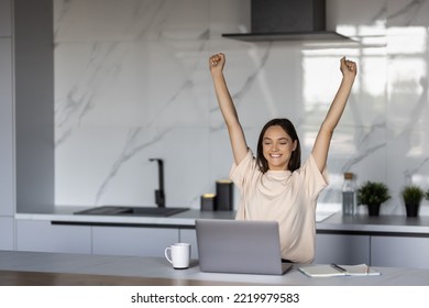 Happy Laptop User Woman Celebrating Good News, Success, Luck With Winner Yes Hands, Shouting For Joy At Laptop In Home Kitchen