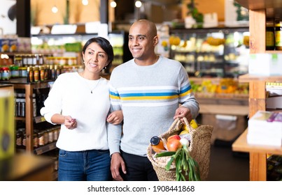 Happy latin-amerikanisches Familienpaar, das in Regalen im Supermarkt spaziert mit Korbbeutel gefüllt mit frischen Lebensmitteln