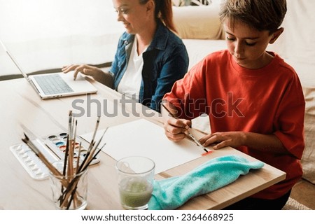 Happy Latin American mother working on computer while child son is painting with watercolors - Home schooling and family love relationship