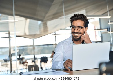 Happy Latin business man wearing glasses working at laptop in office looking away. Happy young male professional using computer sitting at desk thinking of corporate technology solutions. Copy space